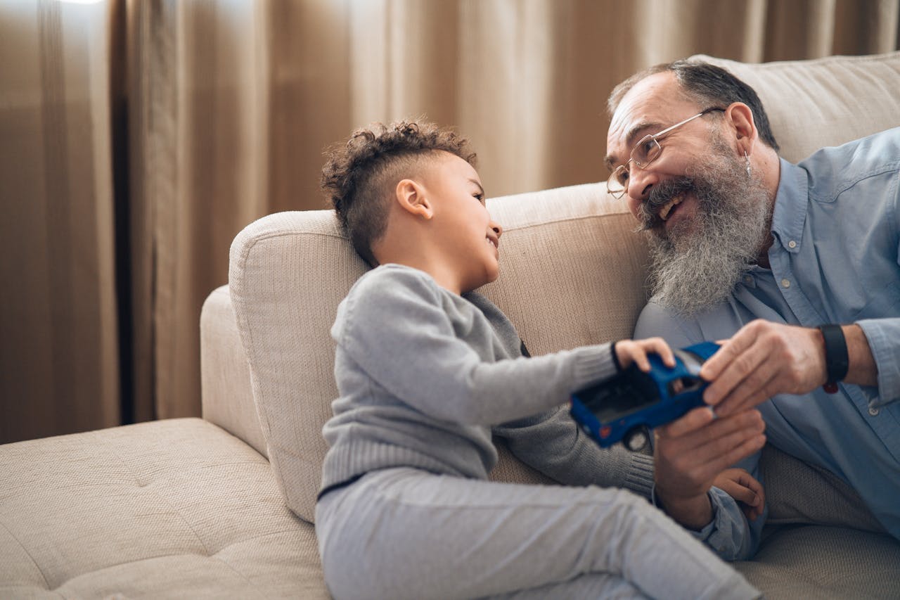 Ein Vater und sein Kind sitzen auf der Couch und schauen sich an. Dabei müssen sie lachen während der Sohn dem Vater ein Spielzeugauto reicht.