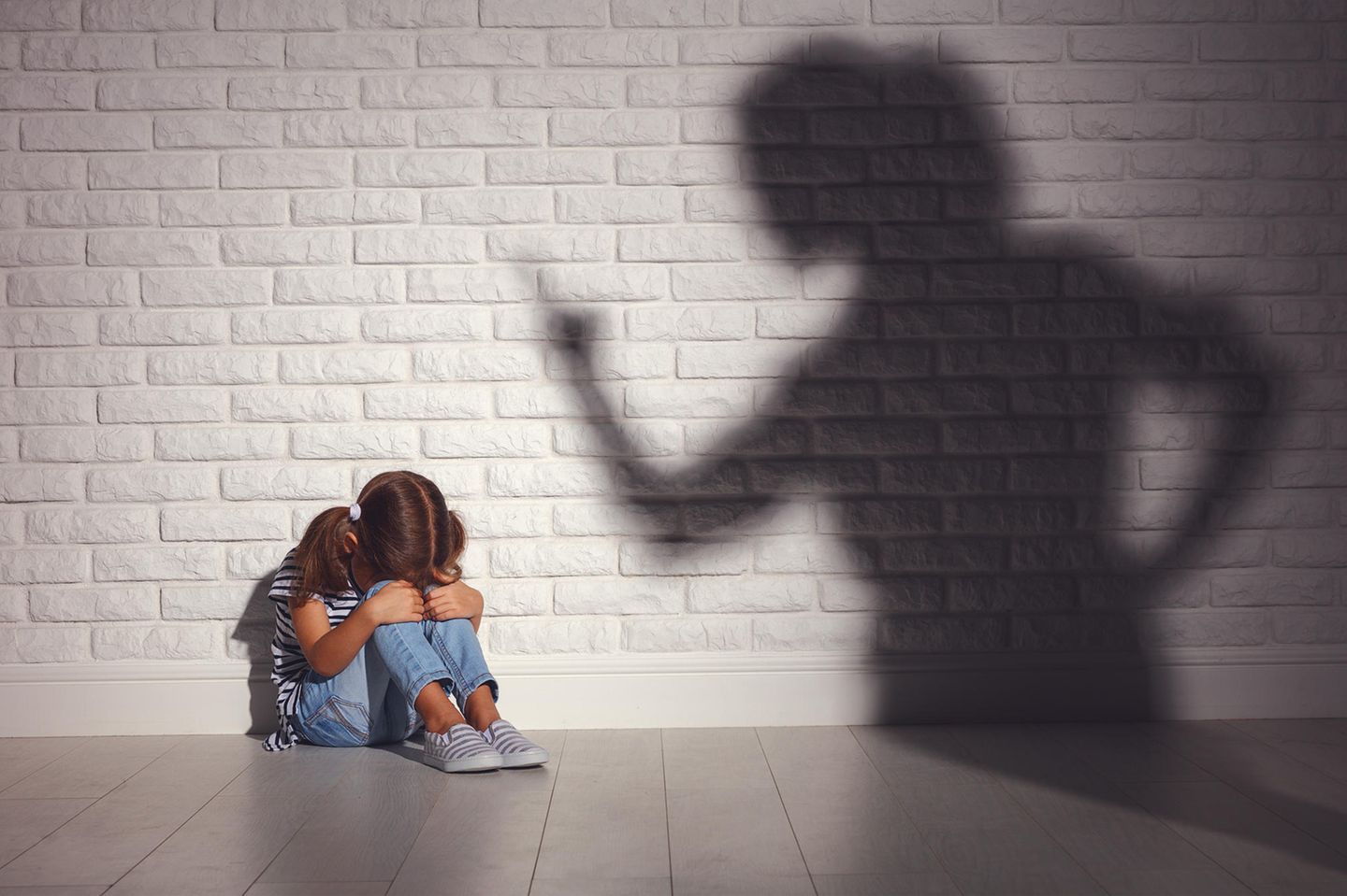 Ein Kind sitzt an der Wand und hat die Arme über den Beinen verschränkt. Der Schatten der Mutter mit erhobenen Zeigefinger wirkt einschüchternd auf das Kind.